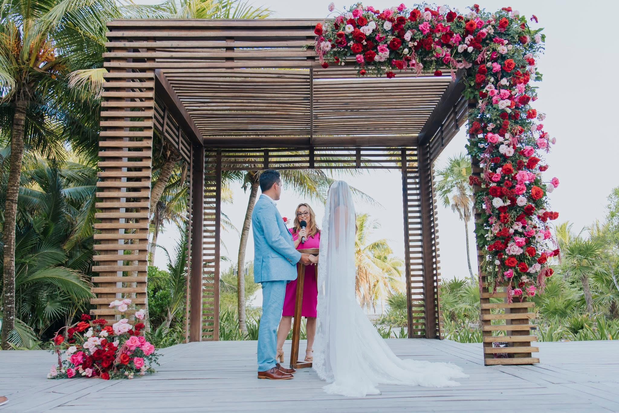 Amy leading the wedding ceremony beneath a pergola decorated with pink and red flowers